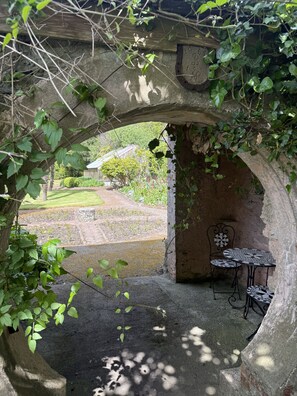 Un arc en pierre abrite une table et une chaise, et mène à un jardin avec une pelouse et un bâtiment en arrière-plan.