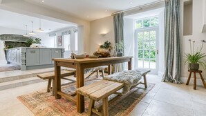 A spacious kitchen with a central island, wooden dining table, and bench, surrounded by large windows and curtains. - Treiorwerth -  a dog that sleeps 15 guests  in 7 bedrooms (Heart of Anglesey)
