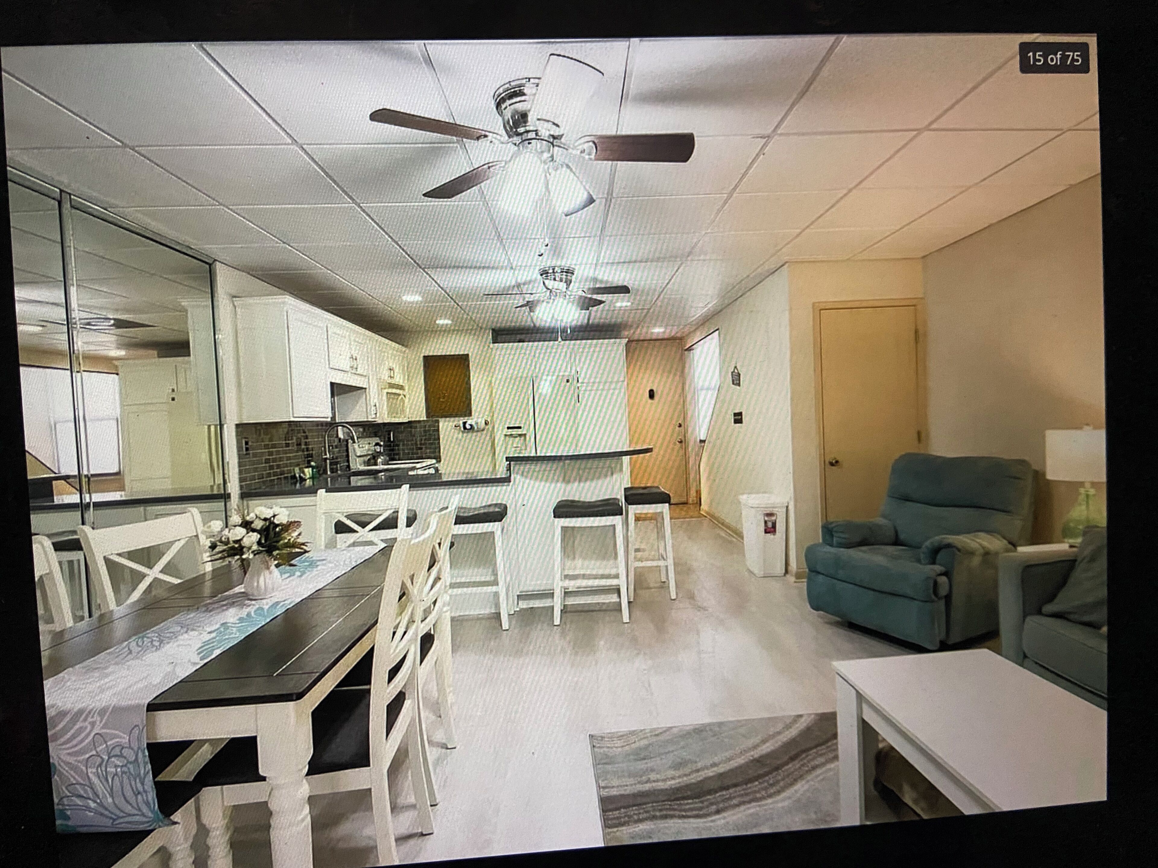 A modern kitchen with white cabinets, a dining area with white chairs, a ceiling fan, and a teal armchair.