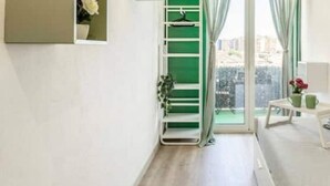 A bedroom with a bed, a small bedside table with a vase and a cup, and a view of a balcony through a window with green curtains.