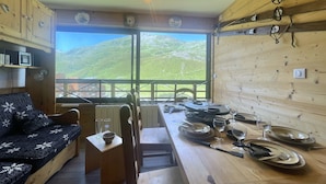 A cozy room with a wooden interior, a sofa, a dining table set with plates, glasses, and cutlery, and a view of green hills through large windows. - (3.4344) Appartement 60m2 sur les pistes 3 Vallées (Les Menuires)