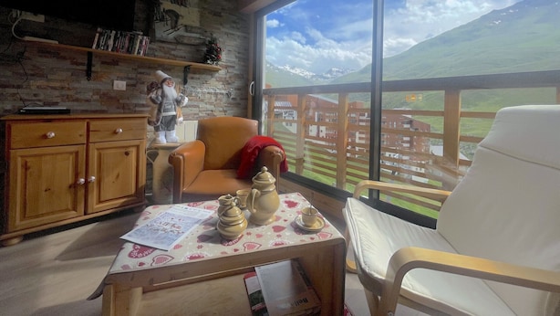 A cozy room with a wooden ceiling, a stone wall, a large window with a mountain view, a wooden cabinet, a table with ceramic items, and a white chair. - (3.4344) Appartement 60m2 sur les pistes 3 Vallées (Les Menuires)