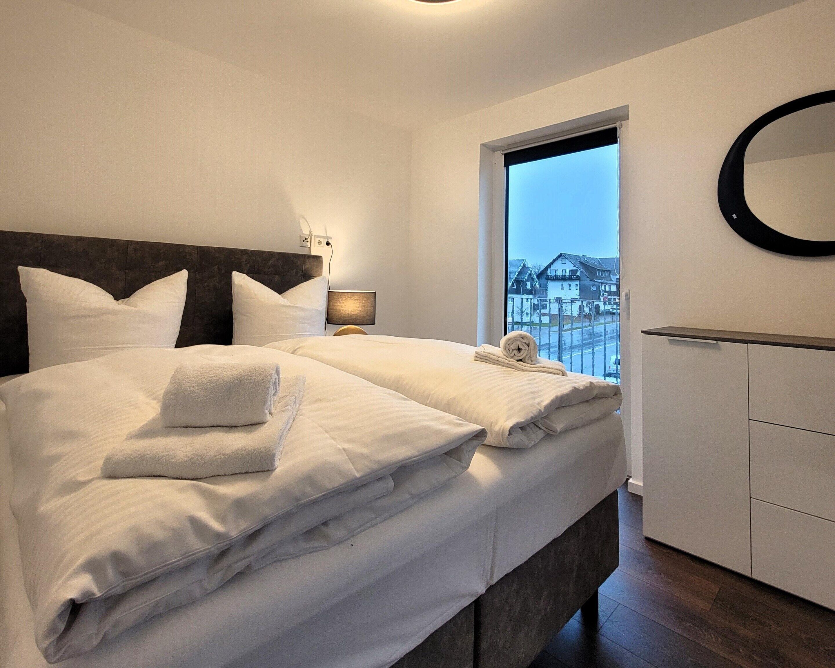 A hotel room with a large bed, a bedside lamp, a window with a view of buildings, and a round mirror on the wall.