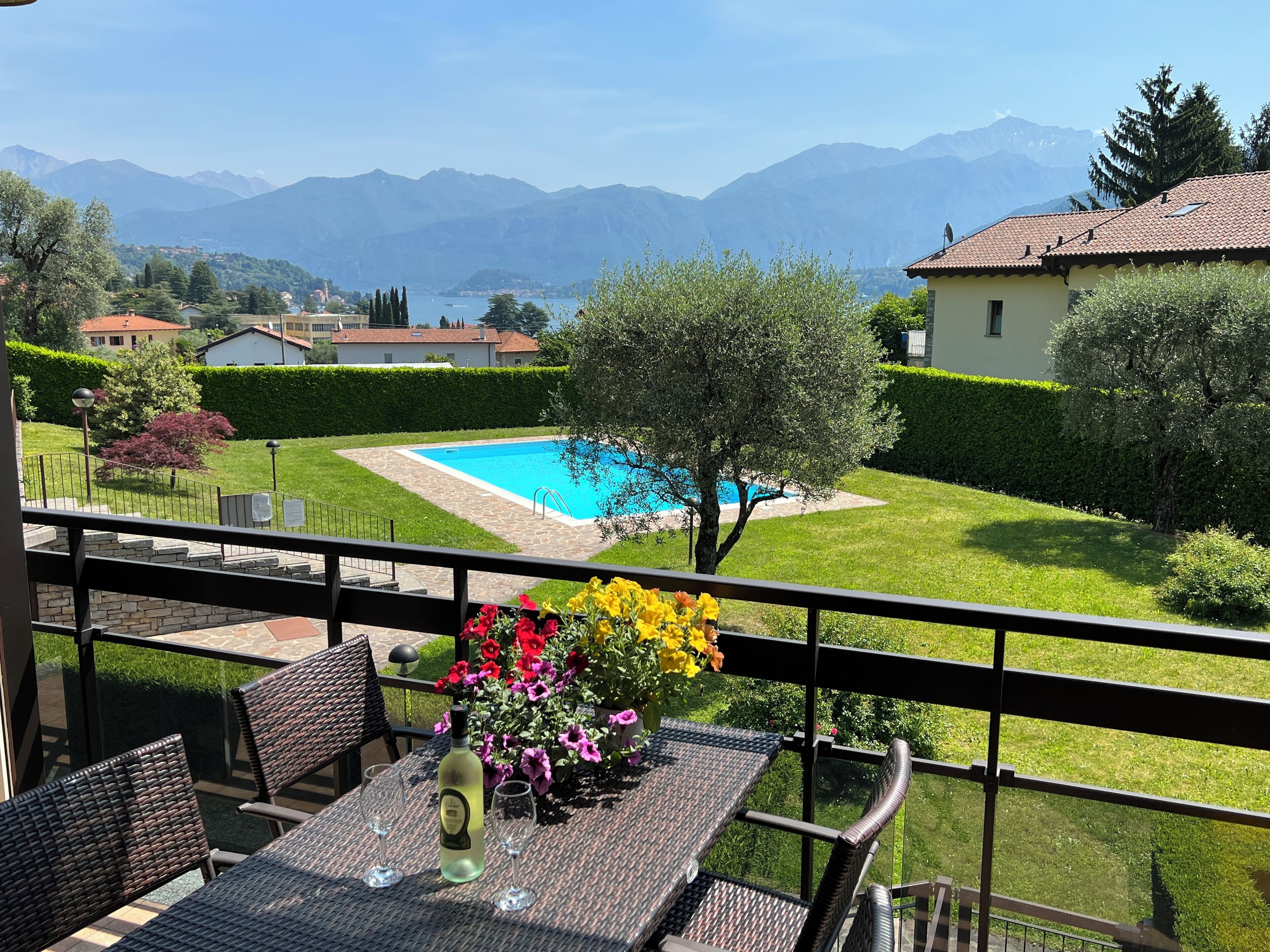 A balcony with wicker furniture, a table with a bottle and glasses, and a view of a swimming pool with mountains in the background.