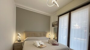 A neatly made bed with a tufted headboard, two bedside tables with lamps, a large window with sheer curtains, and a wall-mounted mirror.