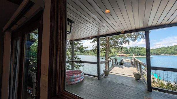 A wooden deck with a view of a lake, a boat, and a dock with chairs and tables. - Smith Mountain Sunrise: Waterfront Party Deck, Great Water Views, & Hot Tub! (Huddleston)