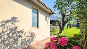 Una casa con terraza de madera, una ventana y flores rosadas en el jardín.
