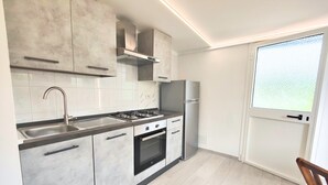 Una cocina moderna con estufa a gas, pileta de acero inoxidable y horno integrado. Se observa un refrigerador a la derecha y un ventanal que proporciona luz natural.