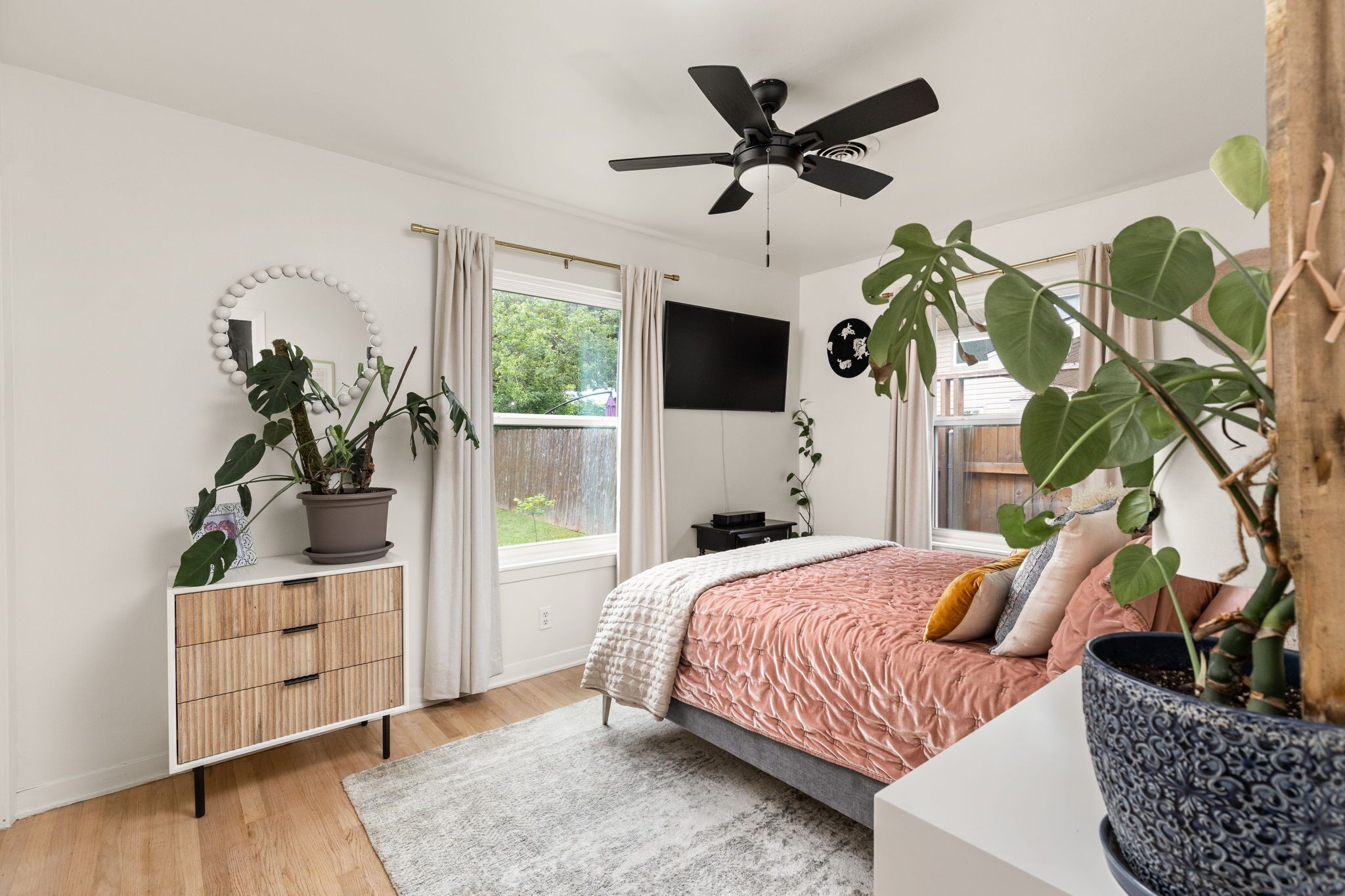 A bedroom with a bed, a ceiling fan, a TV, a window with curtains, a bedside table, and a potted plant.