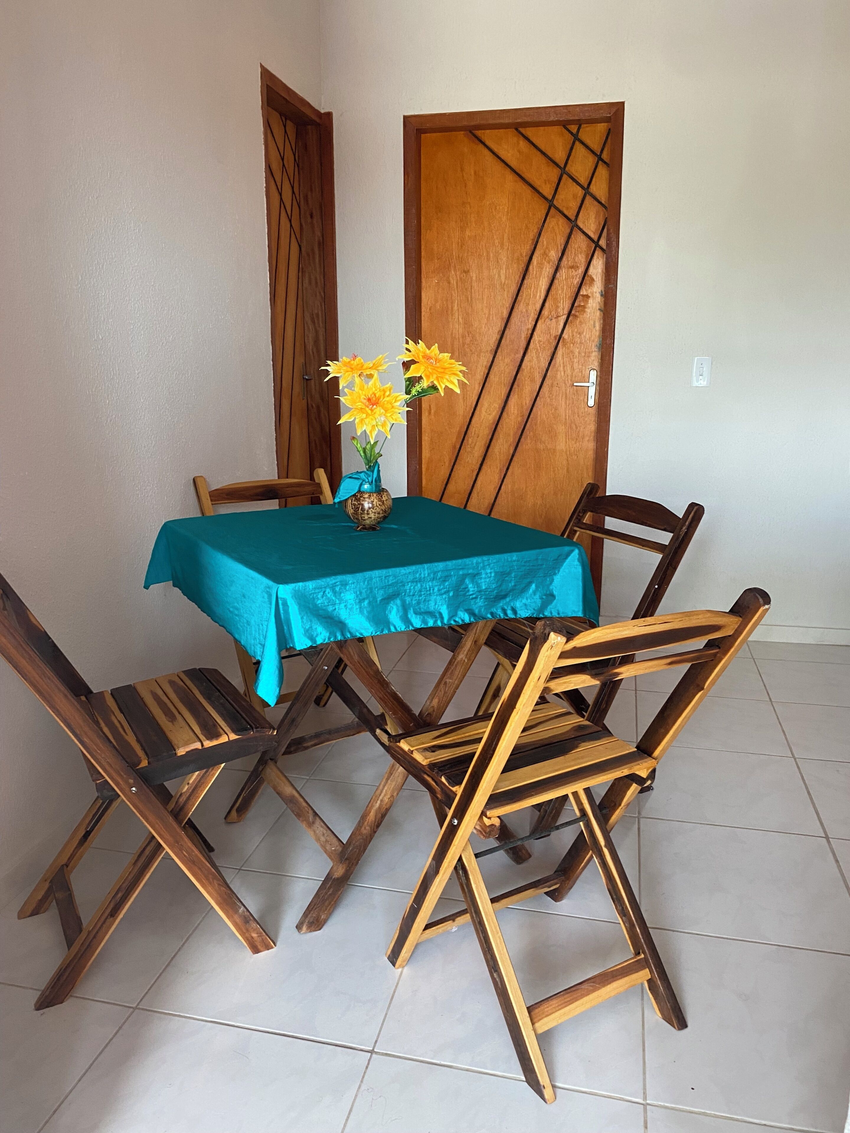 Una mesa de comedor con mantel color turquesa, rodeada de sillas de madera con respaldo en forma de cruz, y un florero con flores amarillas.