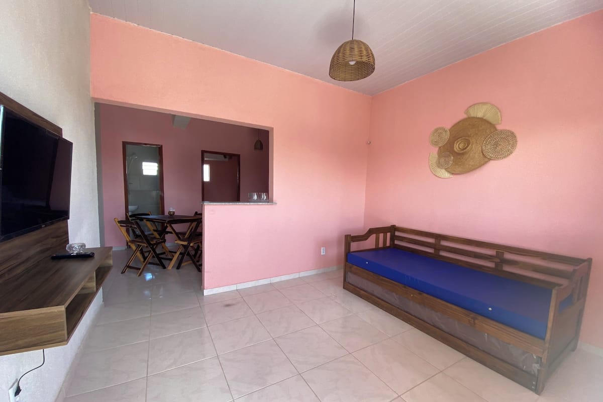 Un salon comprenant un téléviseur, un banc en bois, une table à manger, des chaises et un mur rose agrémenté d’objets décoratifs.