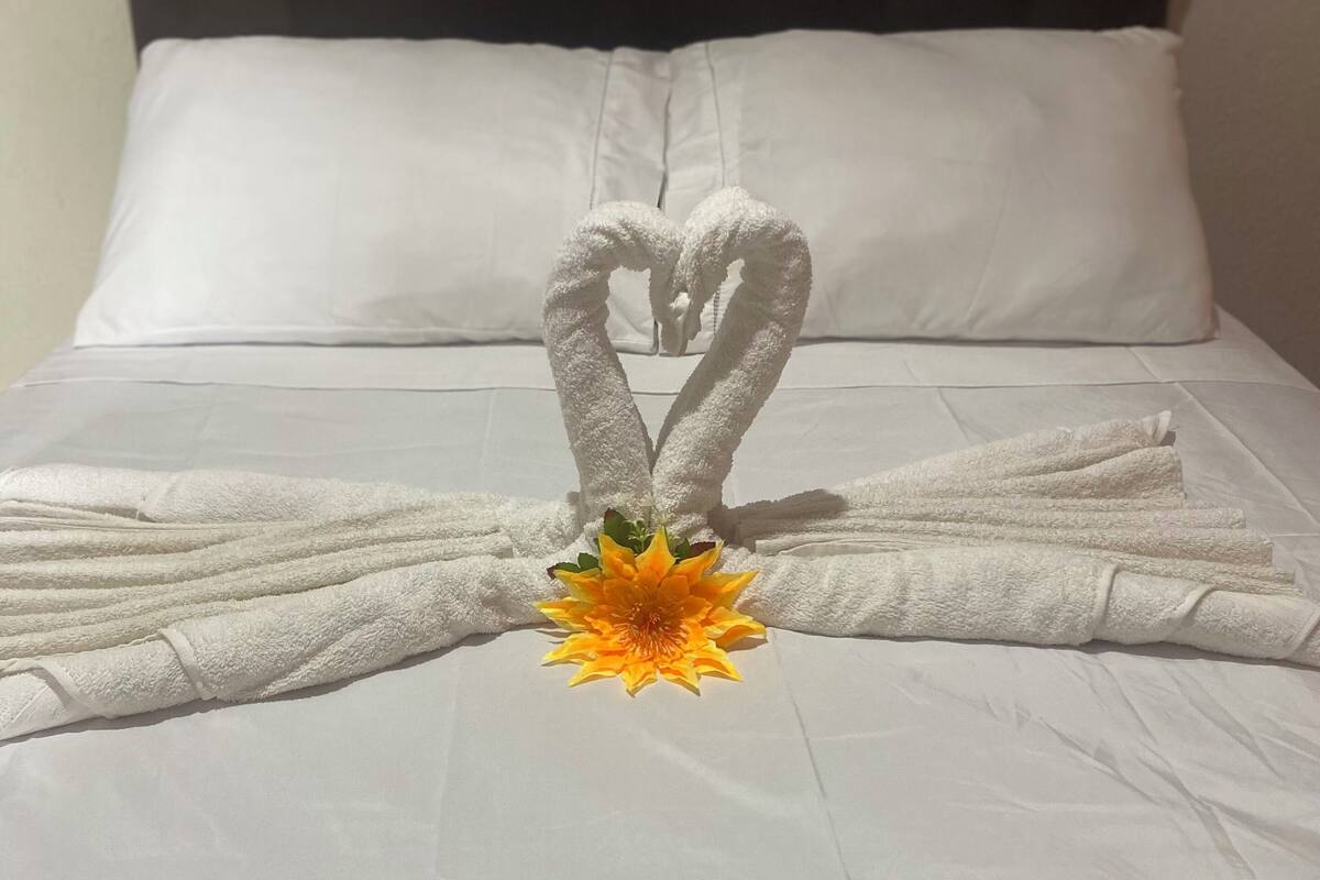Un lit avec du linge de lit blanc, une disposition de serviettes en forme de cœur et un bouquet de fleurs jaunes en guise de décoration centrale.