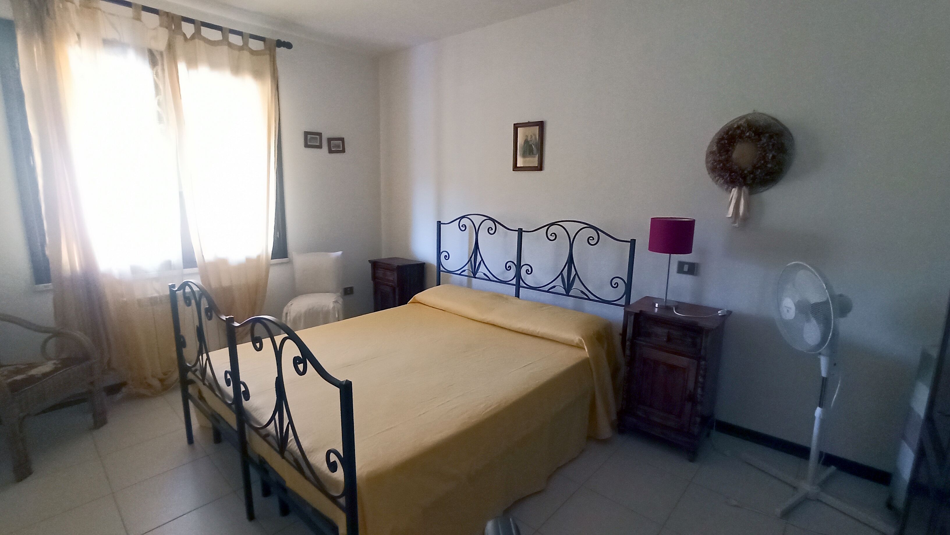 A bedroom with a bed, a chair, a bedside table, a lamp, a fan, and a window with curtains.