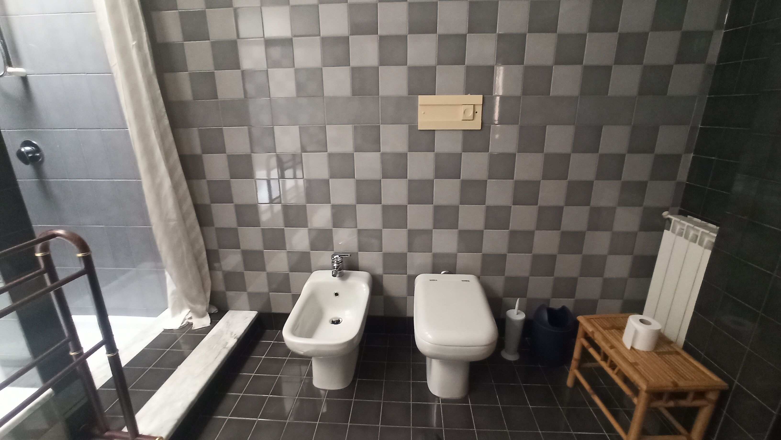 Ein Badezimmer mit Wandfliesen im Schachbrettmuster, einem Bidet, einer Toilette, einem kleinen Holztisch mit Toilettenpapier und einem Handtuchhalter.