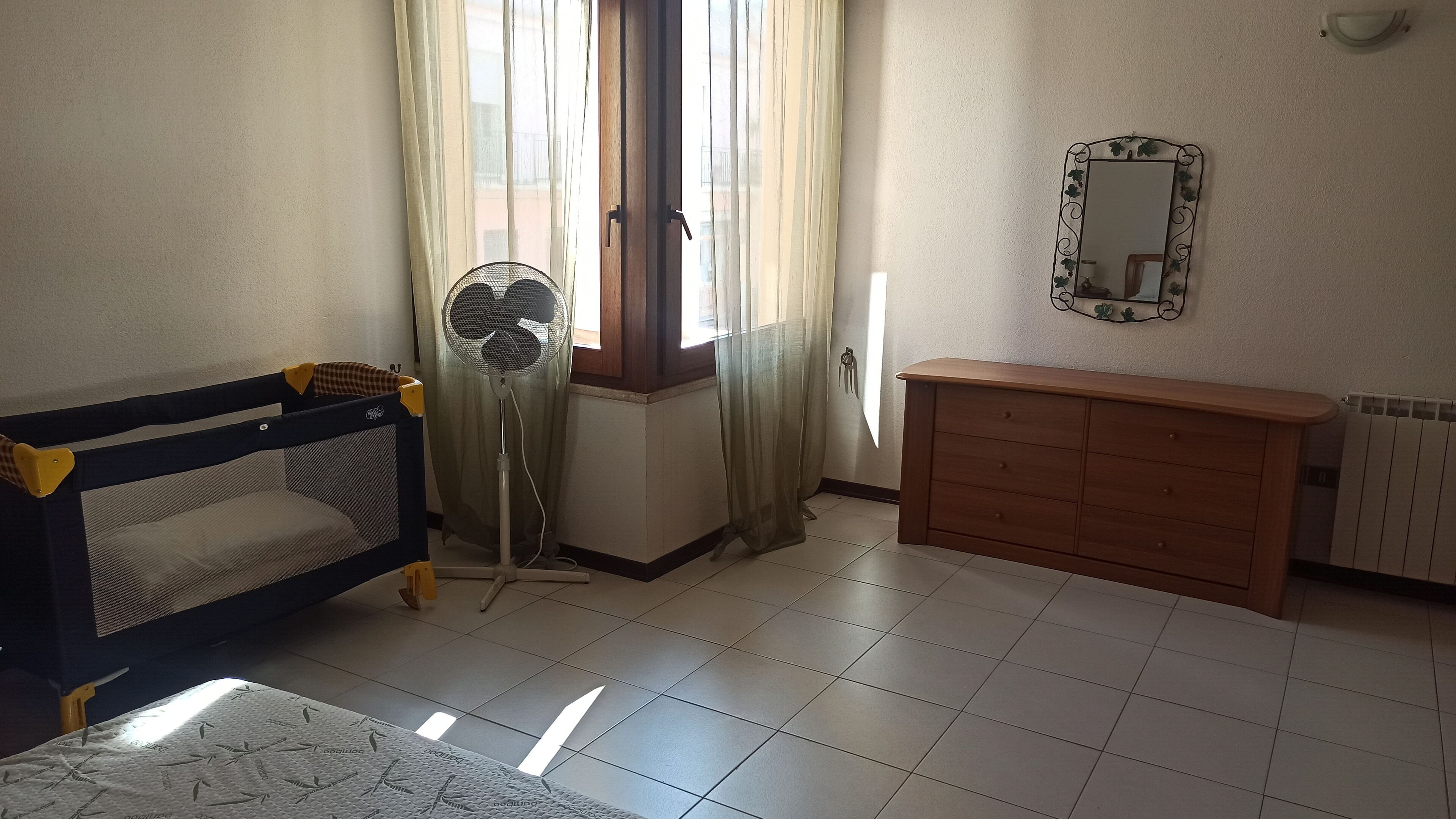 Una habitación con una cuna, un ventilador de pie, un aparador de madera y un espejo en la pared.