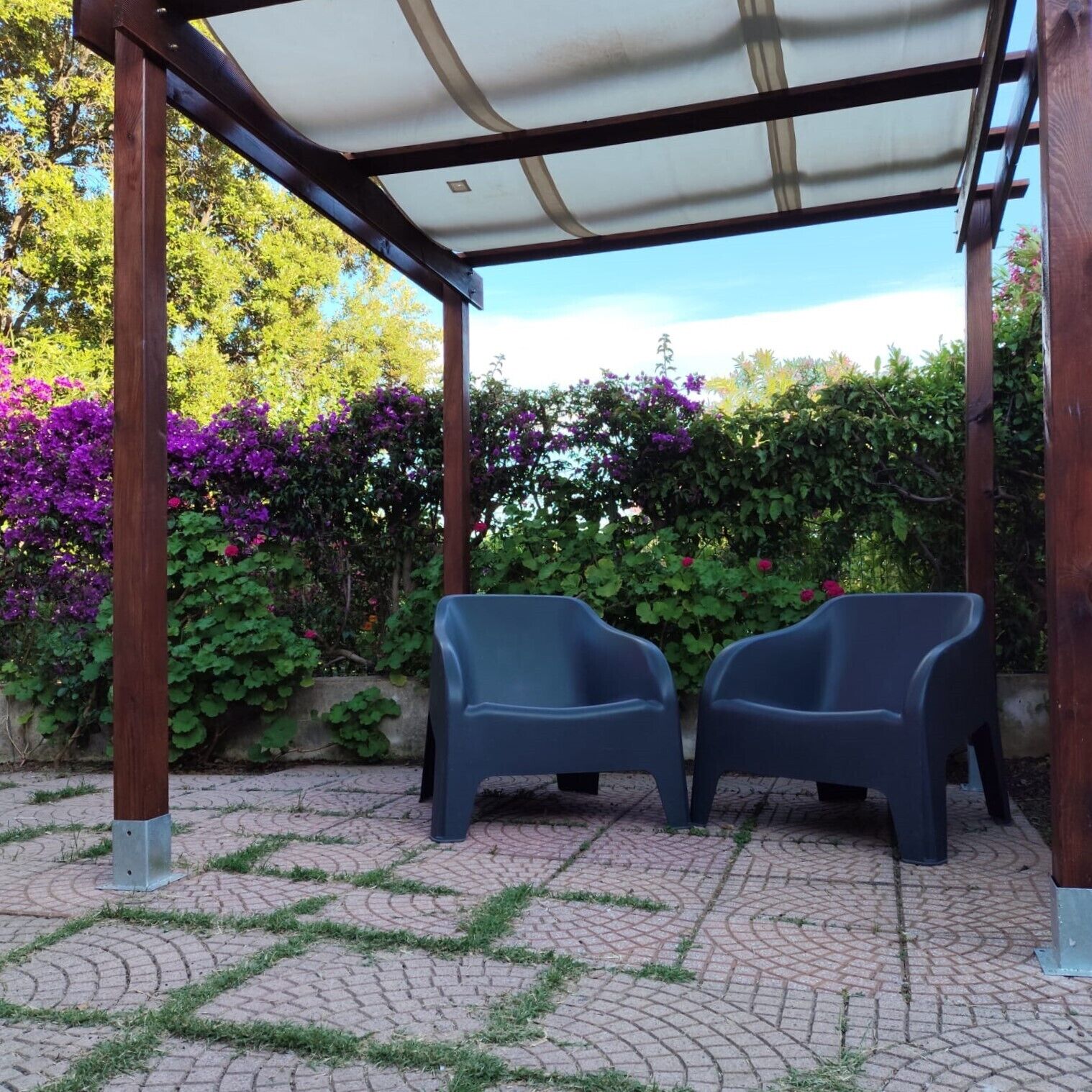 Ein überdachter Außenbereich mit zwei Plastikstühlen, umgeben von einem Garten mit violetten Blüten und grünem Laub.