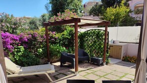 Een terras met een houten pergola, een comfortabele stoel, een klein tafeltje en een tuin met bloeiende planten.