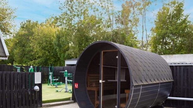 Uma estrutura cilíndrica e moderna com telhado curvo, possivelmente um hotel cápsula ou um alojamento peculiar, situada numa área ajardinada com vedação de madeira e vegetação ao fundo.