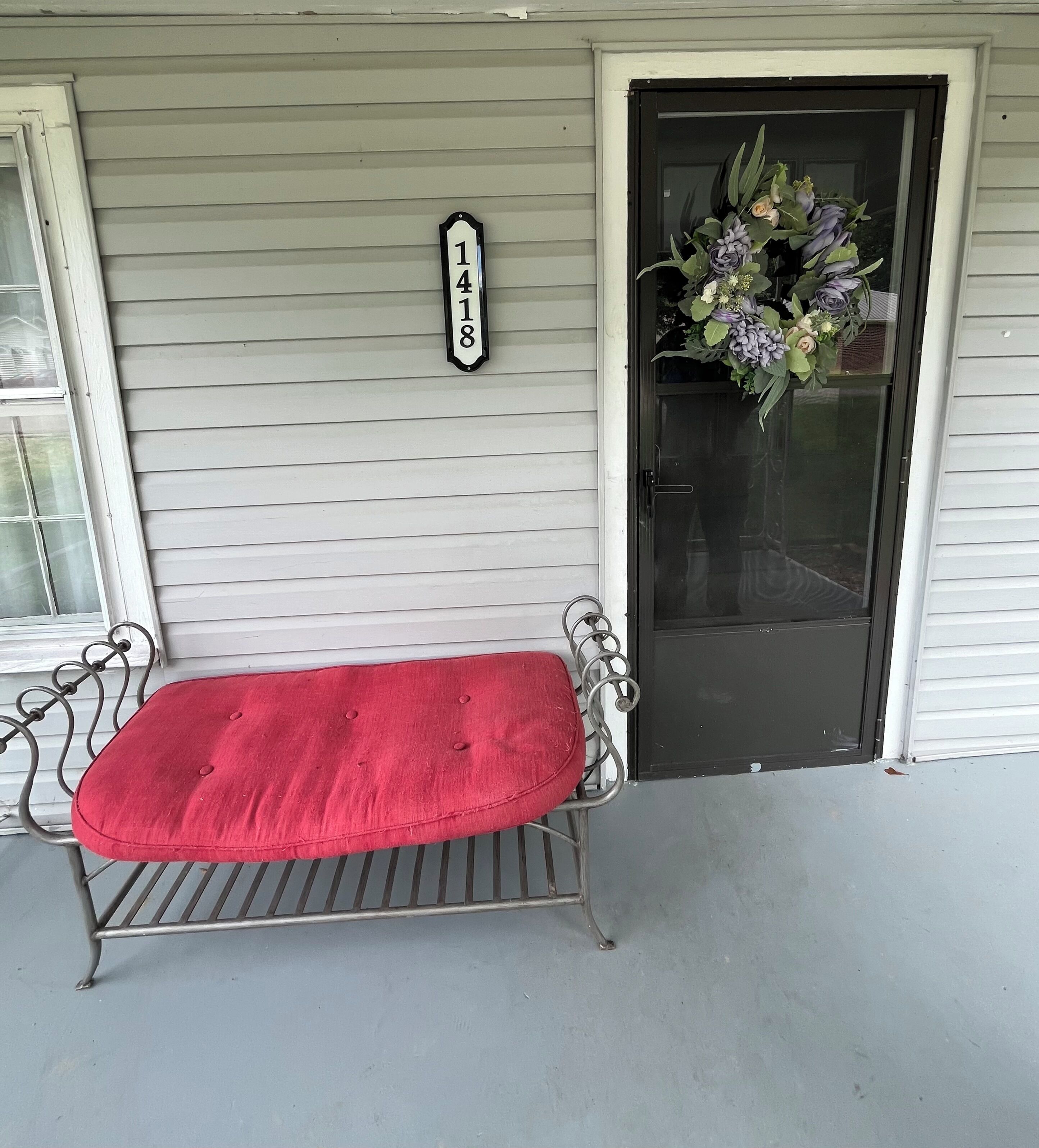 Un banc rouge sur un porche, avec une plaque indiquant le numéro de la maison et une porte ornée d’une guirlande de fleurs.