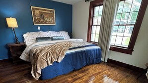 Un dormitorio con suelo de madera, una cama con edredón azul y beige, una mesita de noche con lámpara y un cuadro en la pared.