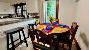 Cocina compacta con armarios blancos, una mesa de comedor de madera con individuales de colores y una ventana con vistas al verdor.