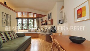 A modern living room with a sofa, wooden dining table, and chairs. There is a window with a view of trees, and shelves with decorative items.