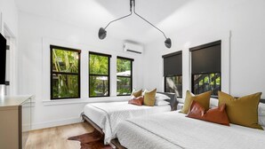 A modern bedroom with two beds, a wooden floor, a cowhide rug, and large windows with a view of greenery. - Hacienda Flamingo Unit 1 - Spacious Designer Flat with Sunroom & Style (West Palm Beach)