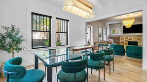 A modern living room with a glass dining table, green upholstered chairs, a large window, and a hanging light fixture. - Hacienda Flamingo Unit 1 - Spacious Designer Flat with Sunroom & Style (West Palm Beach)