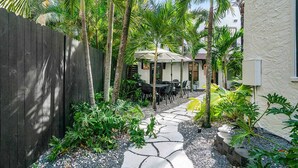 A narrow stone pathway leading to outdoor seating with umbrellas, surrounded by lush tropical plants and a wooden fence.