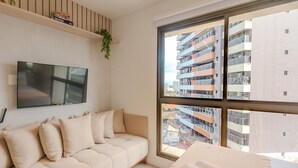 Un soggiorno moderno con un divano, una televisione a schermo piatto, una finestra vista su un edificio residenziale e una piccola pianta su uno scaffale.