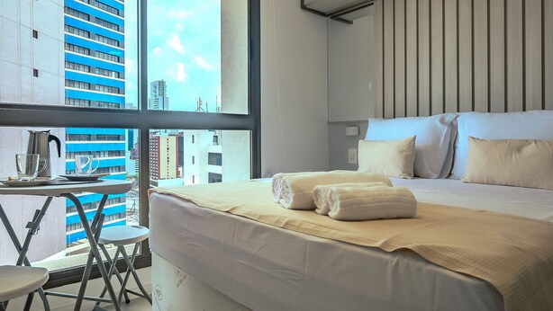 Una camera d'albergo con una grande finestra, un letto con lenzuola bianche, un tavolino con un bollitore e una sedia.