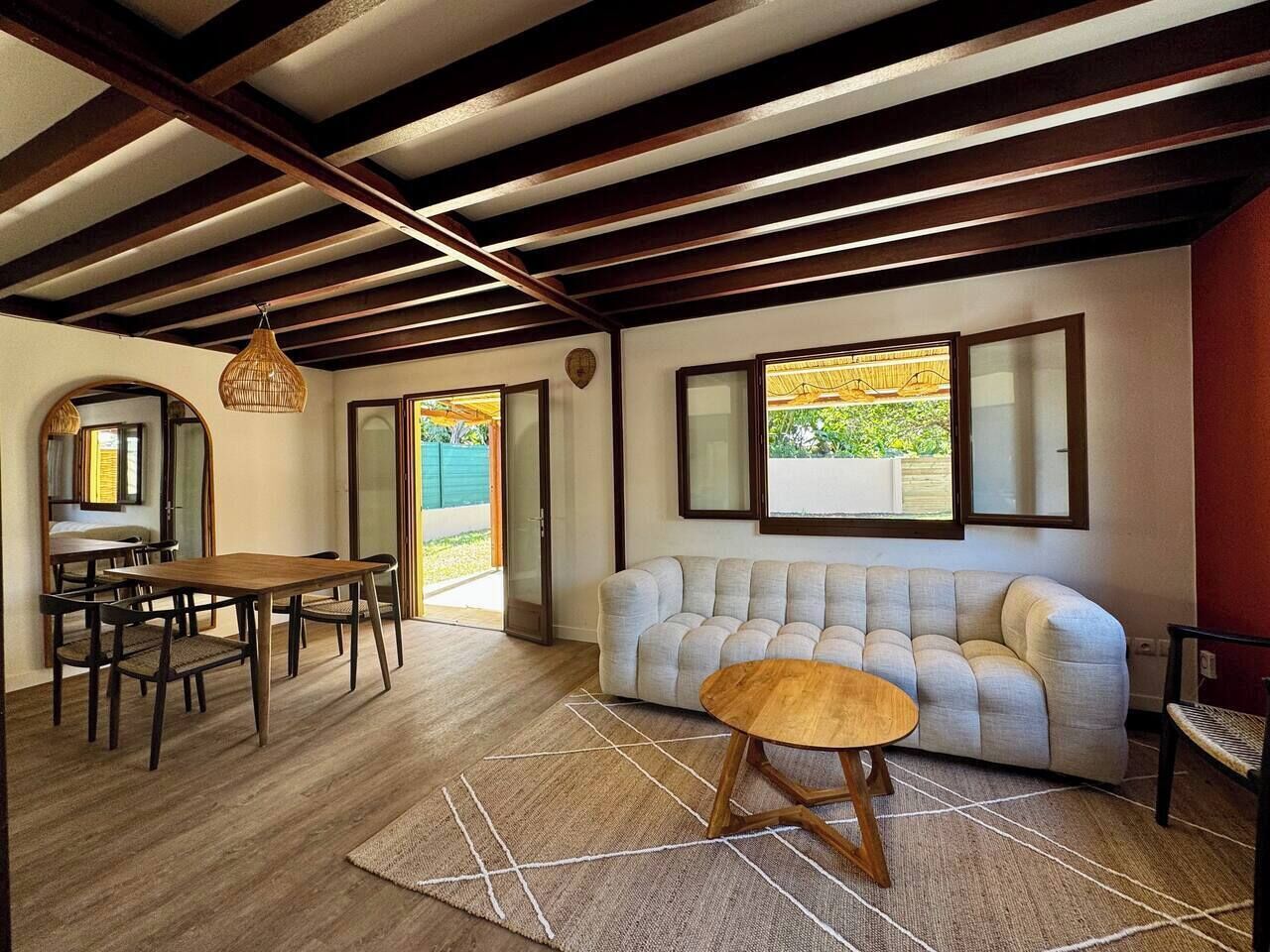 Ampla sala de estar com mesa de jantar de madeira, sofá e mesa de centro. Uma grande janela oferece luz natural e vista para o exterior.