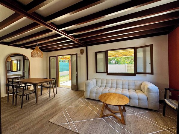 A spacious living area with a wooden dining table, a sofa, and a coffee table. There is a large window providing natural light and a view of the outdoors.