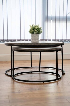 A round table with a black metal frame and a white pot with green plants on top, placed on a wooden floor in front of a window with vertical blinds. - Sevenstay Fleet Apt8 (Merseyside)