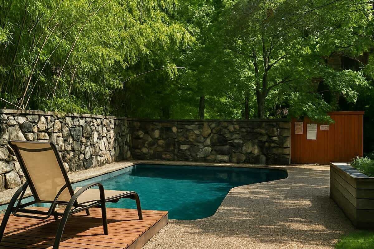 Una piscina con muelle de madera y una tumbona, rodeada por un muro de piedra y abundante vegetación.