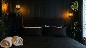 A dark-themed bedroom with a large bed, two bedside lamps, and a potted plant on the left.