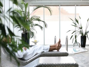 Une personne allongée sur une chaise longue dans une pièce bien éclairée, dotée de grandes fenêtres, et entourée de plantes en pot.