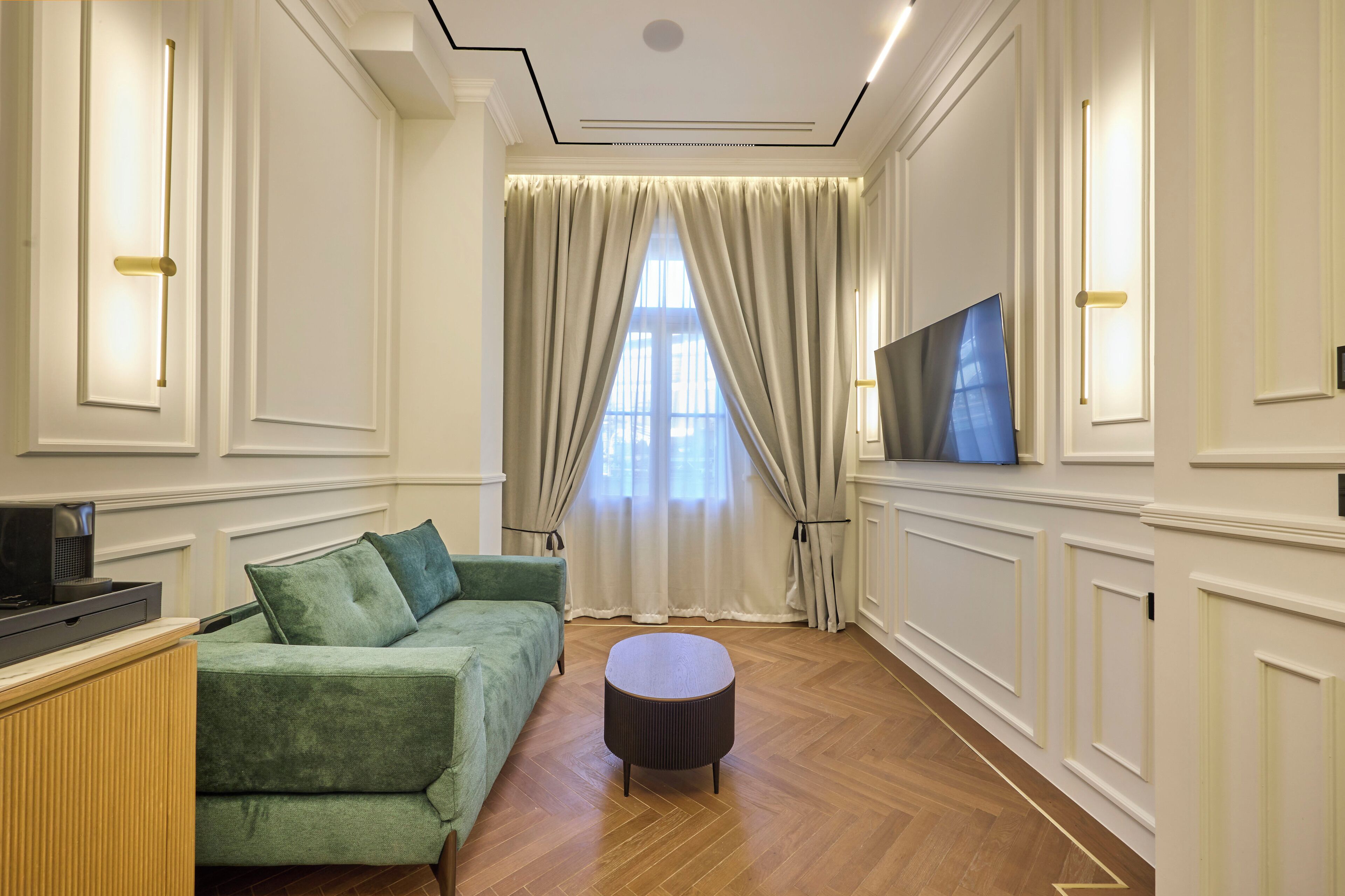 Ein Zimmer mit einem grünen Sofa, einem runden Hocker, einem Flachbildfernseher und einem großen Fenster mit Vorhängen.