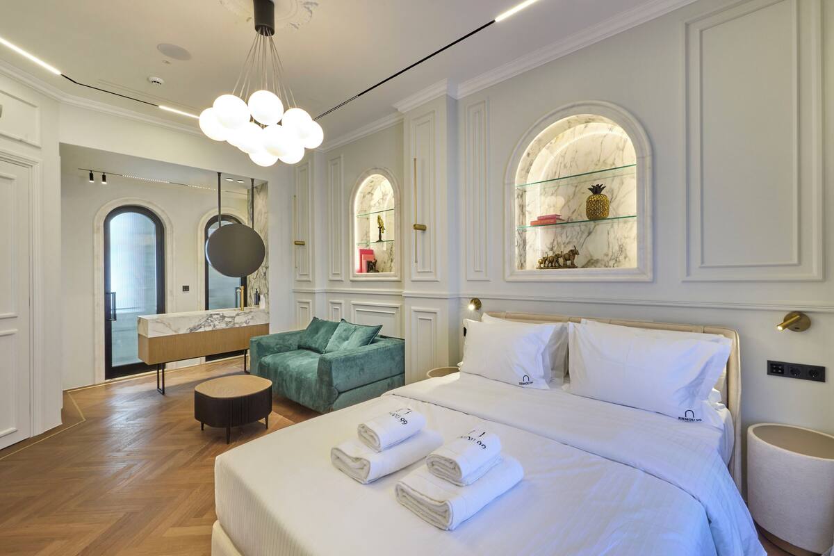 A modern hotel room with a large bed, a green armchair, a marble vanity, and arched windows with shelves.