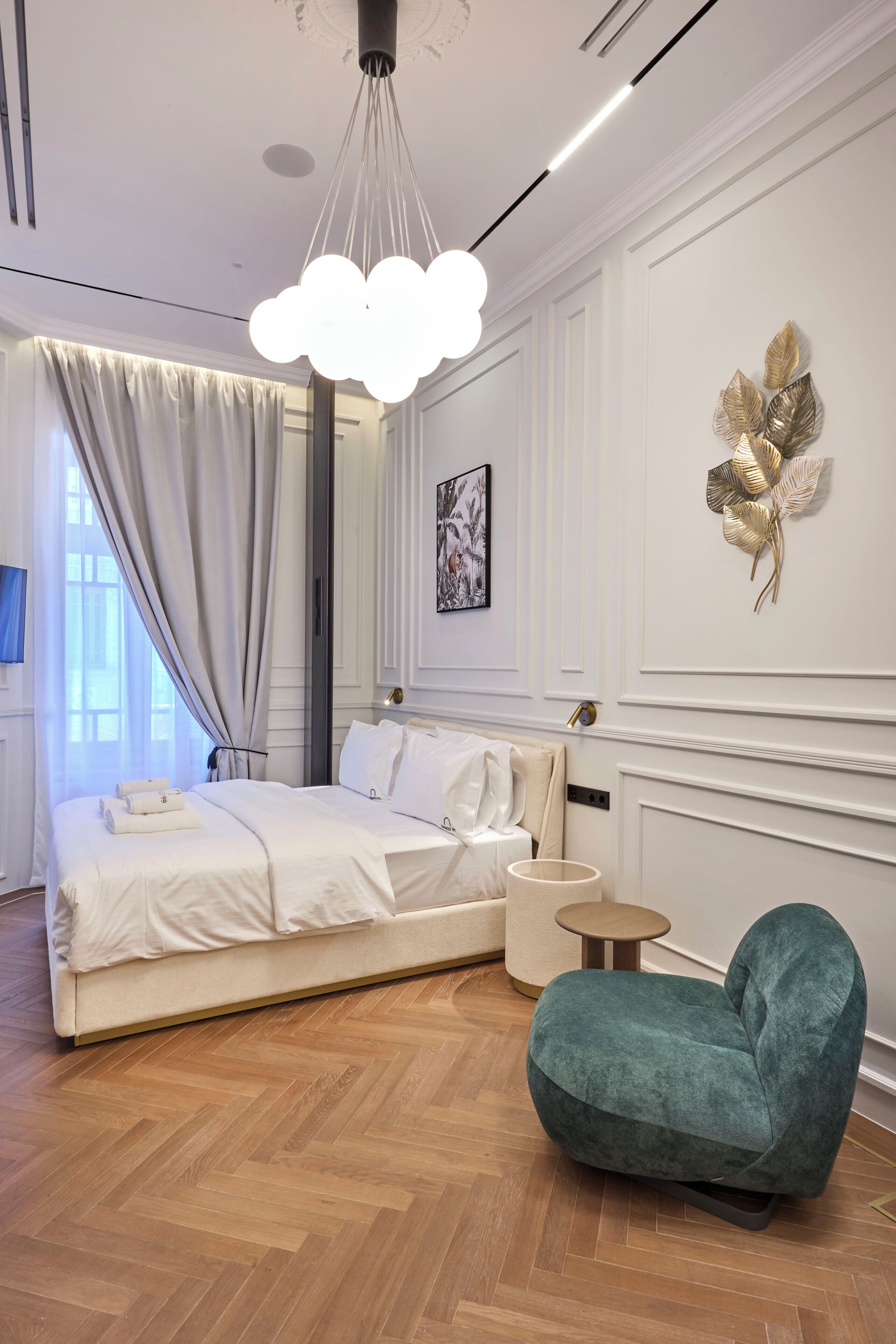 A modern hotel room with a large bed, a teal armchair, a round side table, and a decorative wall piece.