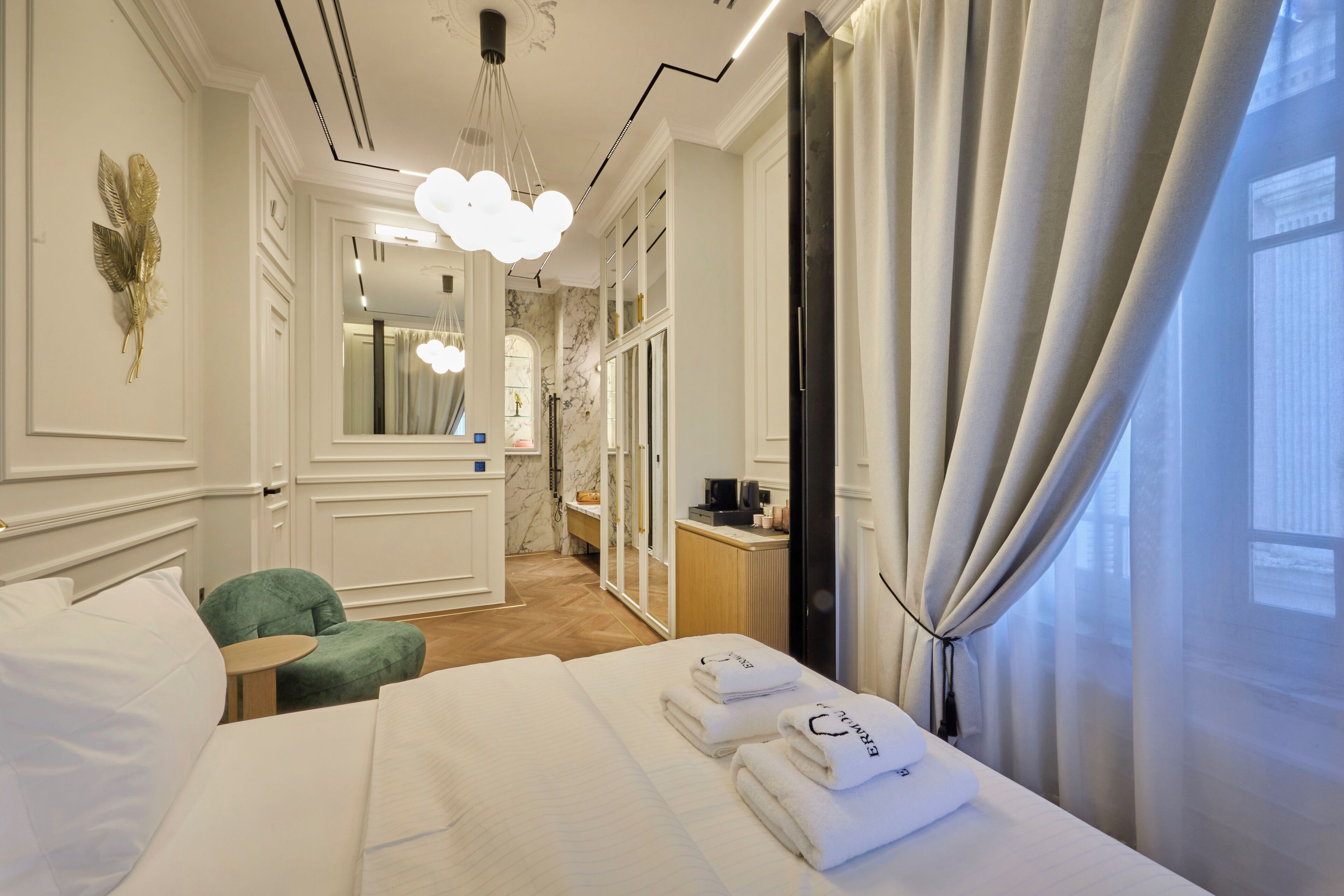 Habitación de hotel con una cama bien hecha, un sillón verde, una mesita y un baño visible a través de una puerta abierta.