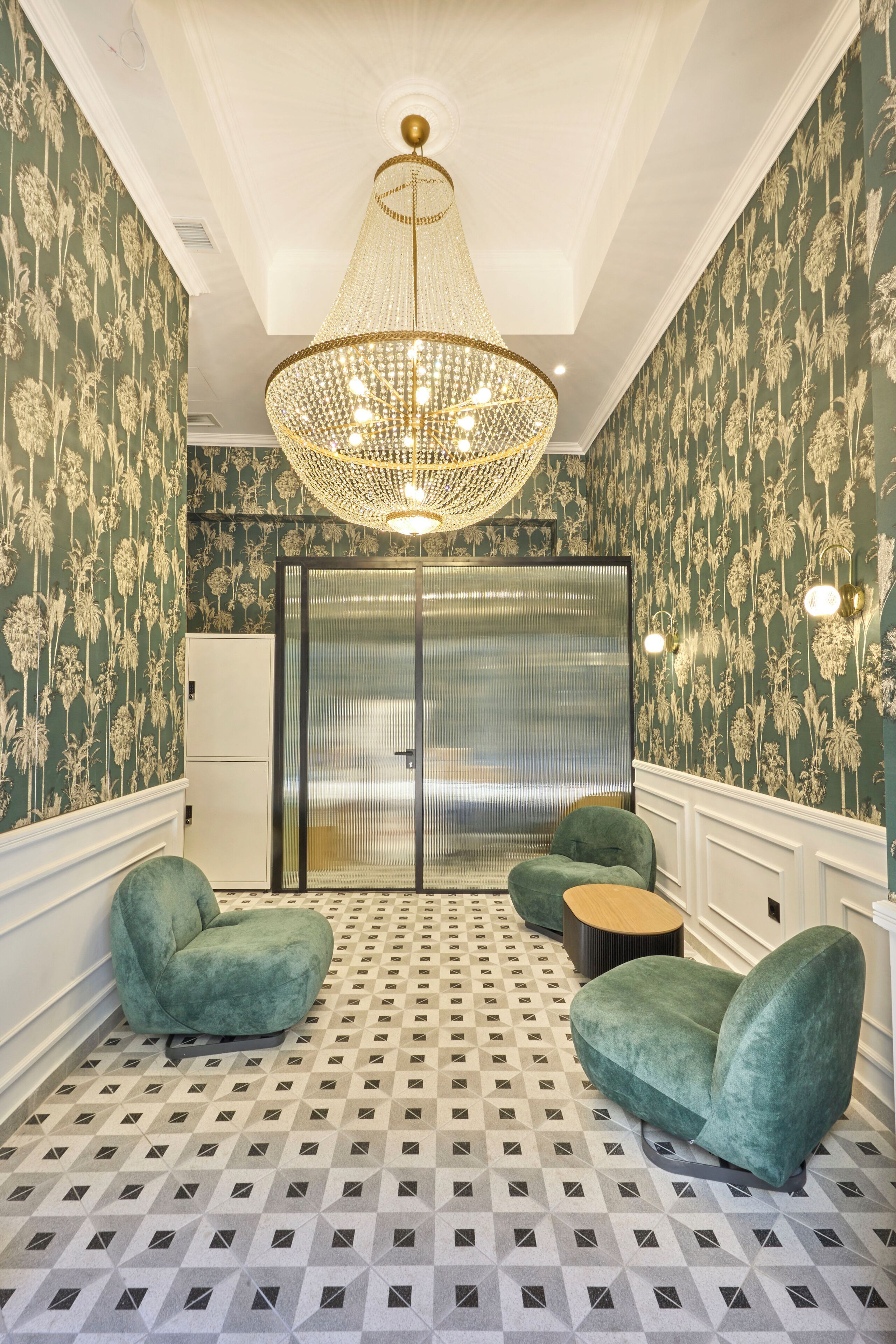 A room with patterned wallpaper, a chandelier, two green armchairs, a small table, and a door with a frosted glass panel.