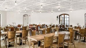 A spacious dining area with multiple wooden tables and chairs, a tiled floor, and a white ceiling with grid patterns.