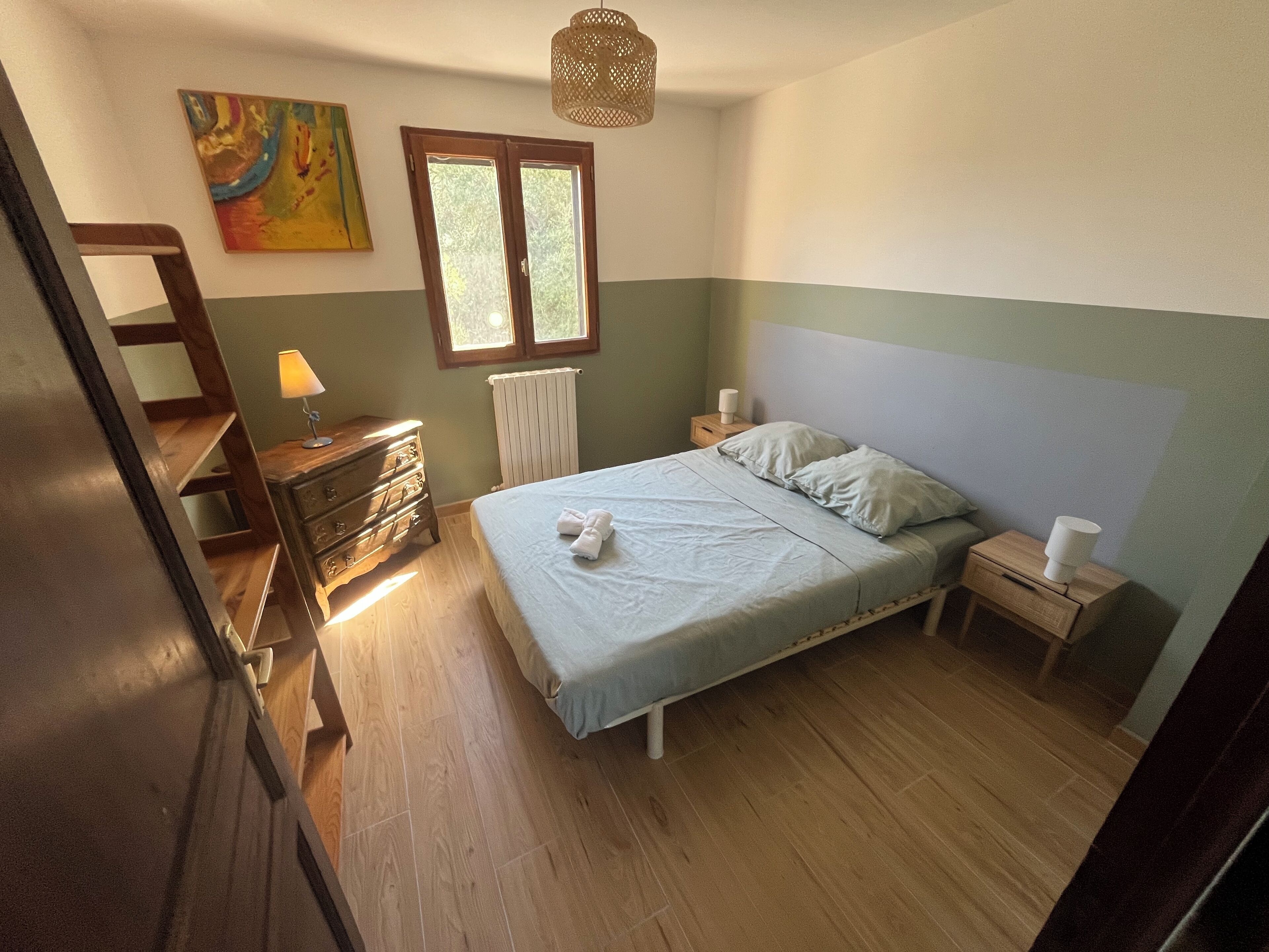 Ein Schlafzimmer mit Bett, Kommode, Nachttisch, Heizkörper, Fenster und einem Bild an der Wand.