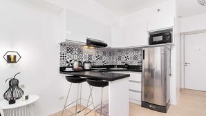 Een moderne keuken met witte kasten, een zwart aanrecht, een roestvrijstalen koelkast en een magnetron. Er staan twee zwarte barkrukken bij een klein wit tafeltje met een kaars.