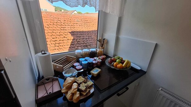 Ontbijt met brood, fruit en verschillende soorten beleg op een tafel bij een raam met uitzicht op een gebouw met rode daken.