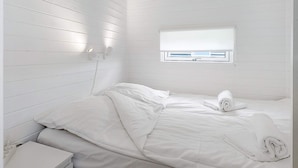 A white room with a bed, a small bedside table, and a window with blinds. - 6 Person Holiday Park Home in Bogense (Bogense)