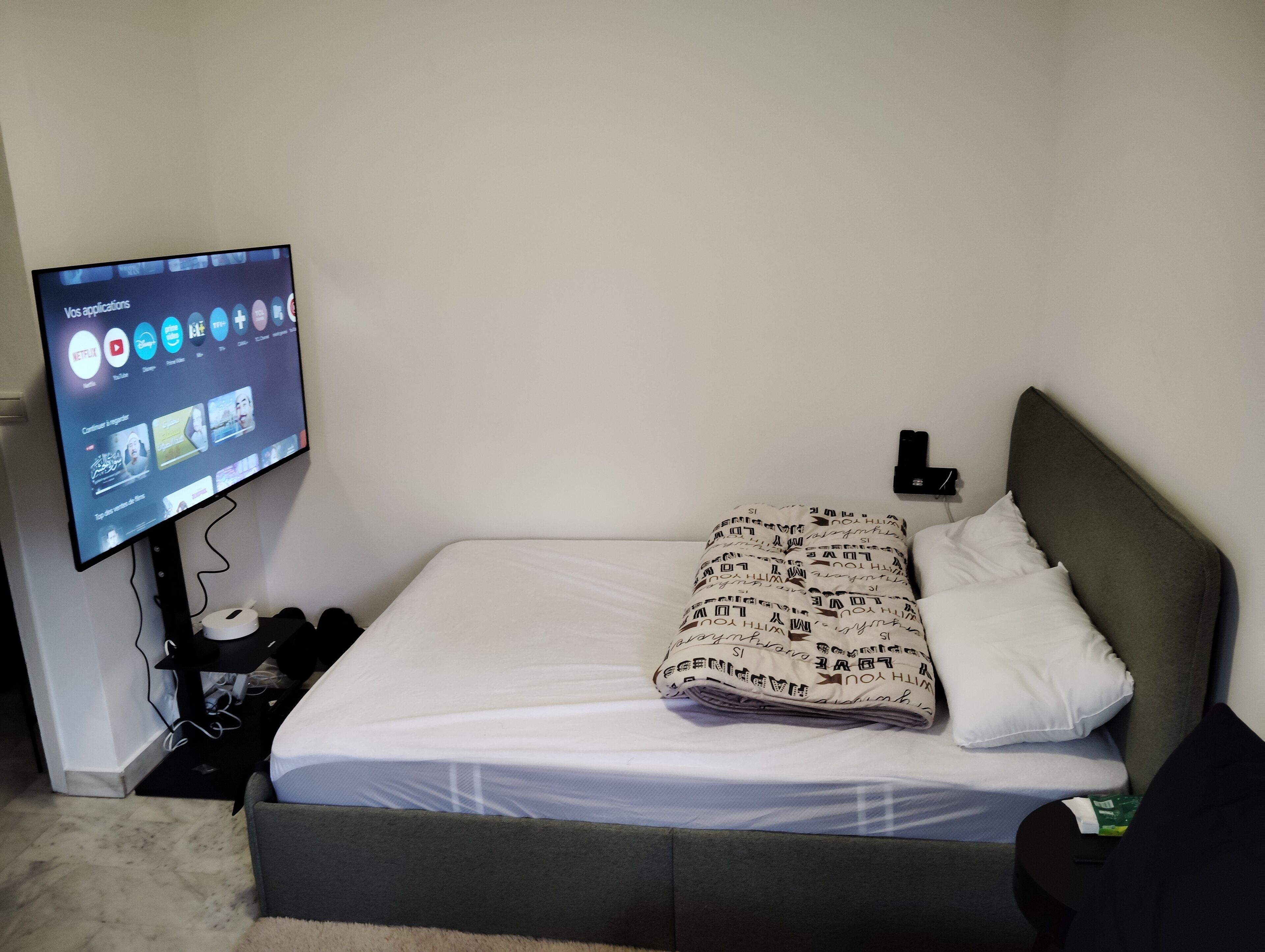 Een net opgemaakt bed met een bedekker en kussens, een flatscreen-tv aan de muur met een streamingdienst interface, en een klein nachtkastje met een telefoon en een groen voorwerp erop.