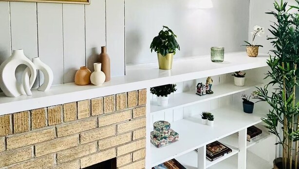 Un salon avec une cheminée en briques, des étagères blanches ornées d’objets décoratifs et un tableau encadré accroché au mur.