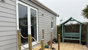 A house with a grey exterior, a wooden front door, a small window, a wooden fence, a green pergola, and a paved patio.